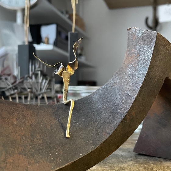 Kleine goldfarbene Figur mit erhobenen Armen auf einem grossen gebogenen Metallobjekt in der Werkstatt