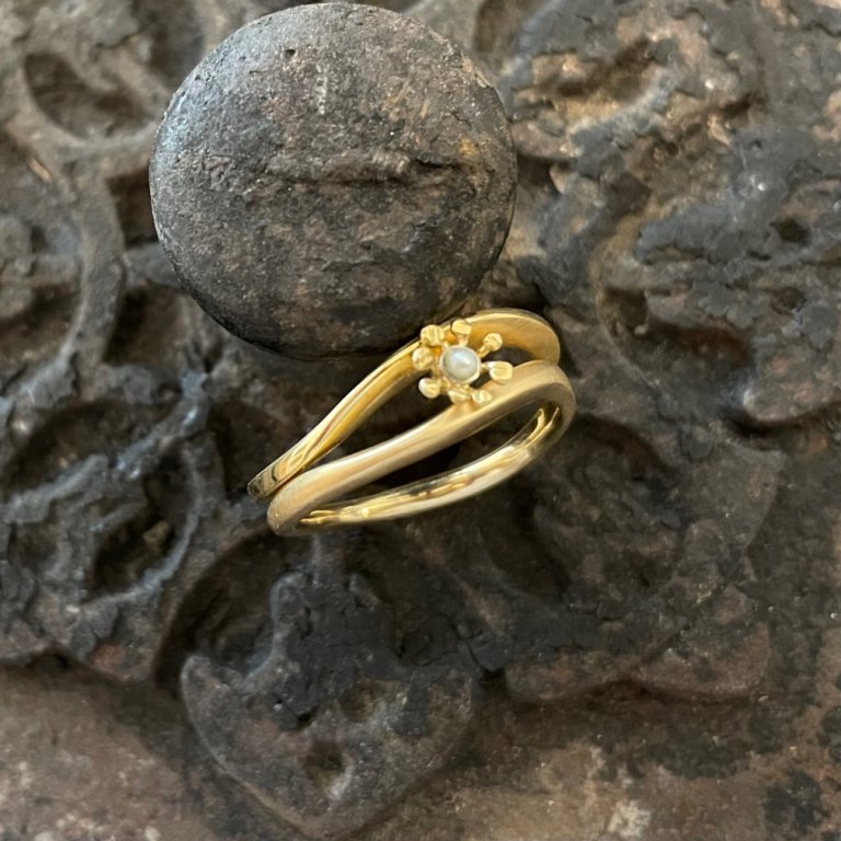 Goldfarbener Ring mit zwei geschwungenen Schienen und kleiner Blüte auf dunklem Metall