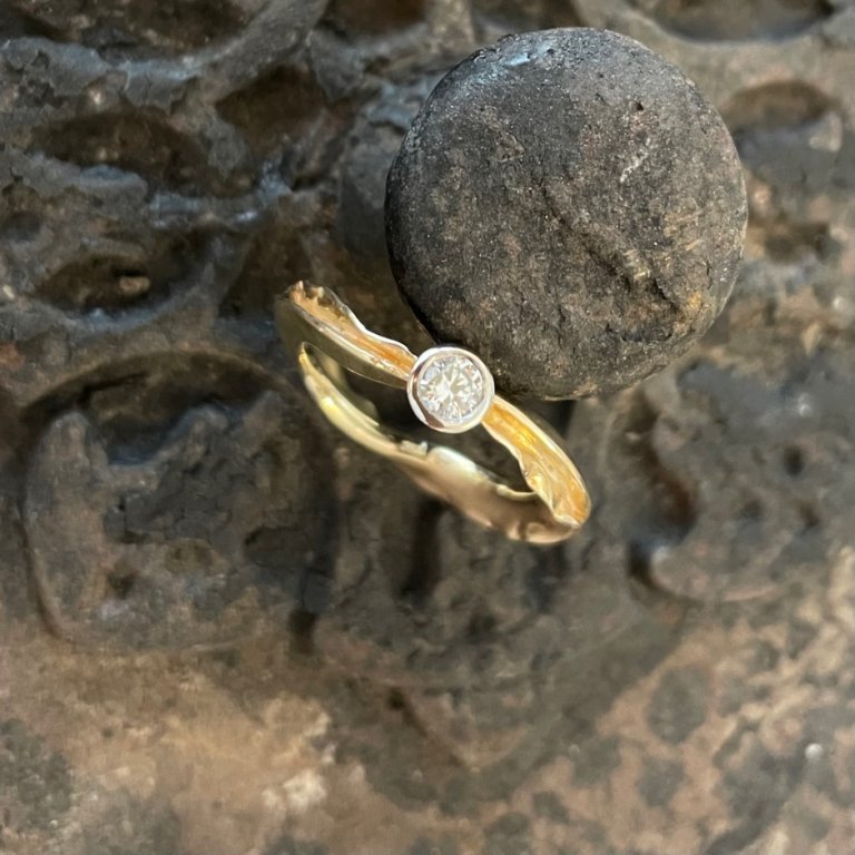Goldfarbener Ring mit geschwungener Ringschiene und rundem hellem Stein auf dunklem Metall