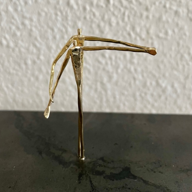 Zwei filigrane goldfarbene Figuren stehen eng hintereinander und strecken jeweils einen Arm seitlich aus.