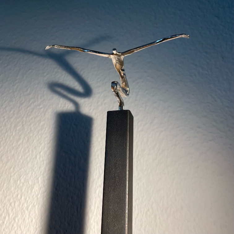 Filigrane Figur mit ausgebreiteten Armen auf hohem dunklem Sockel und markantem Wandschatten