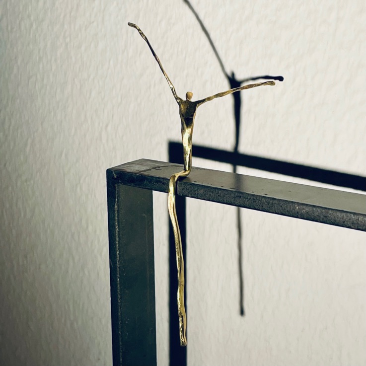 Schlanke Metallfigur mit erhobenen Armen und langem herabhängendem Bein auf einem dunklen Metallrahmen