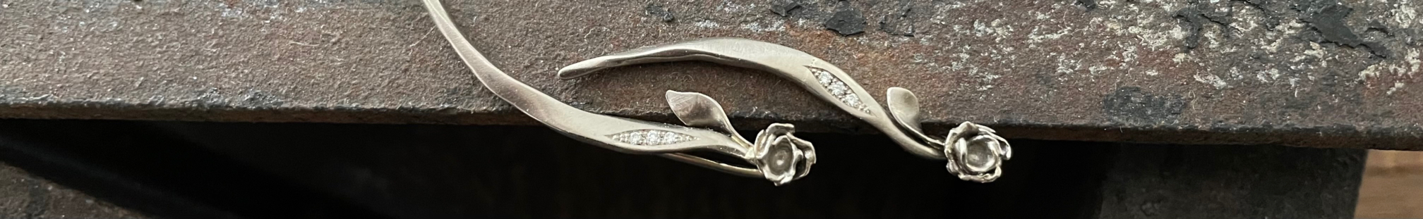 Zwei silberfarbene Ohrringe mit kleinen Rosen, langen geschwungenen Blättern und hellen Steinen auf verwittertem Metall.