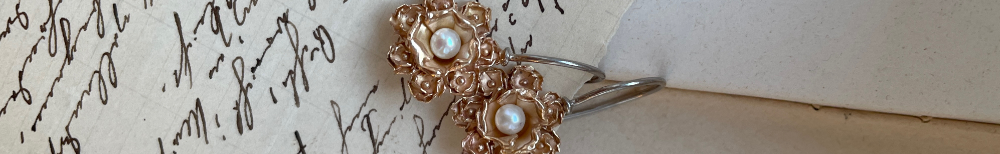 Zwei rotgoldfarbene Ohrringe mit kleinen Blüten rund um eine weisse Perle auf handbeschriebenem Papier.