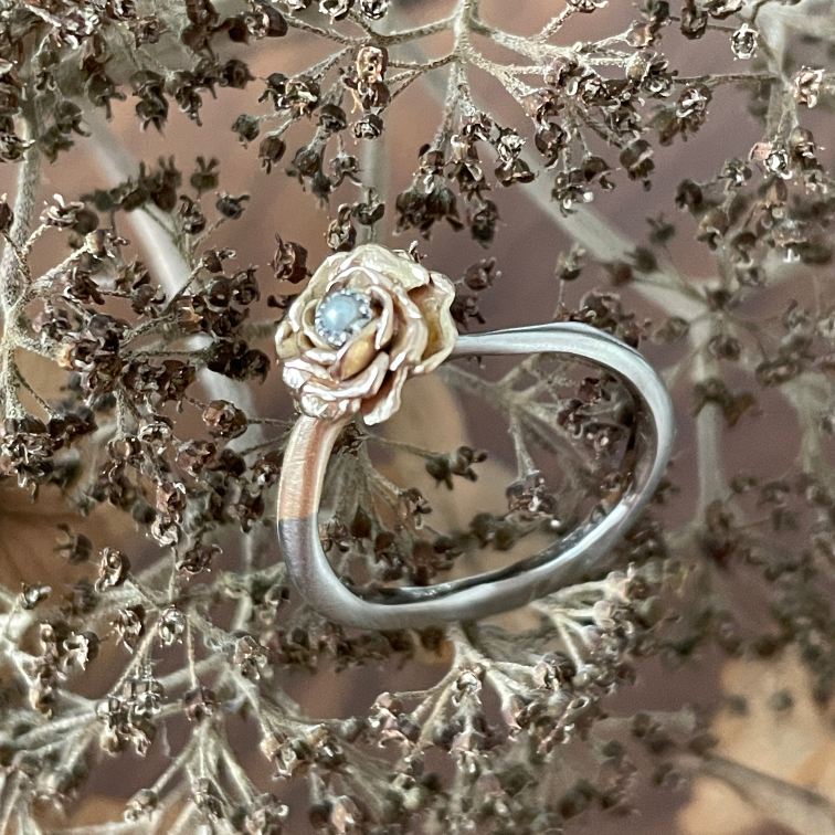 Silberfarbener Anhänger in offener Tropfenform mit goldfarbener Rose und hellblauem Stein zwischen Zweigen