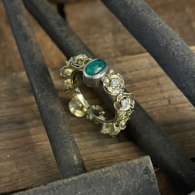 Goldfarbener Ring mit ovalem grünem Stein, kleinen Blüten und hellen Steinen zwischen Goldschmiedewerkzeugen.