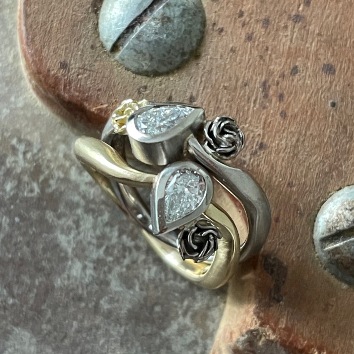Zwei gold- und silberfarbene Ringe mit geschwungenen Ringschienen, dunklen Rosen und klaren tropfenförmigen Steinen auf einem Holzwerkzeug.