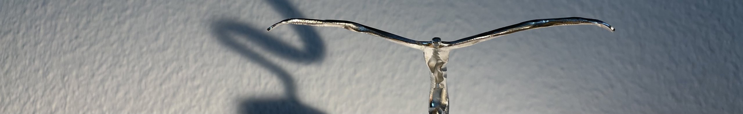 Nahansicht einer filigranen Metallfigur mit ausgebreiteten Armen und ihrem Schatten an der Wand.